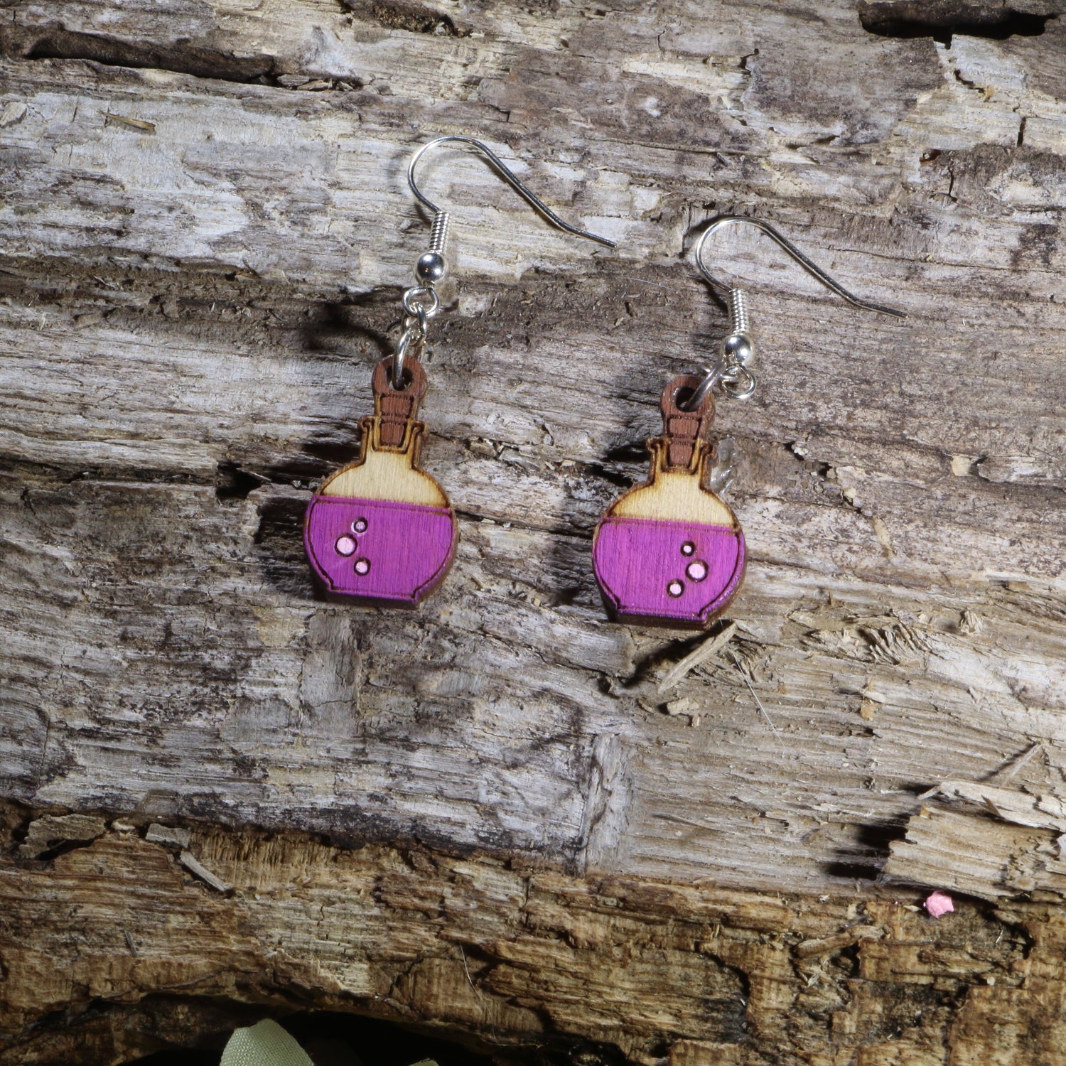 Handcrafted wooden potion bottle dangle earrings featuring vibrant purple potion details and engraved accents. These lightweight fantasy-inspired earrings are finished with hypoallergenic .925 sterling silver hooks, making them the perfect accessory for gamers, fantasy fans, witches, alchemists, tabletop RPG players, and lovers of magical jewelry.