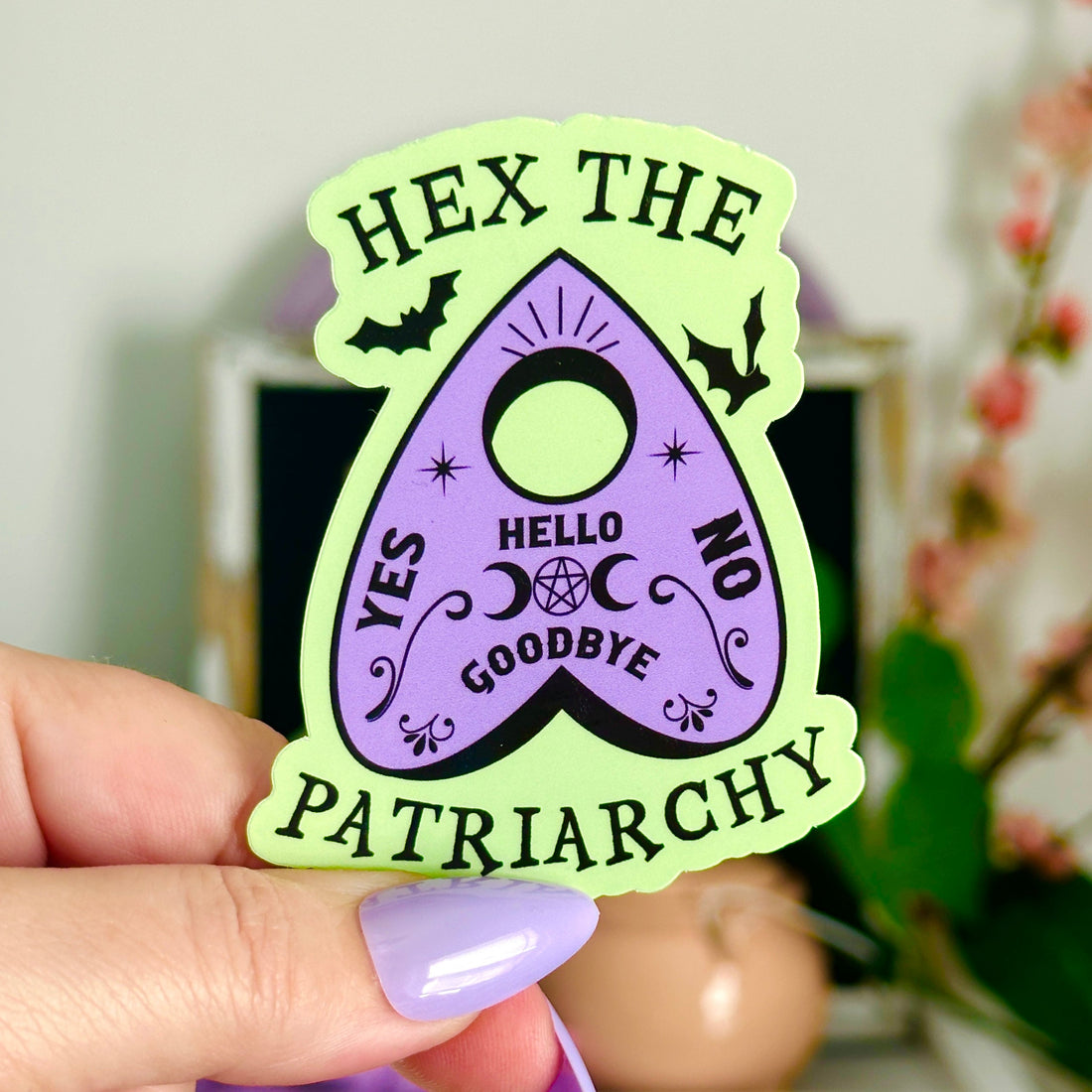 Hex the Patriarchy witchy planchette sticker in pastel green and lavender with bats, moons, and stars, feminist witchcraft laptop decal resting on a lavender skeleton hand.