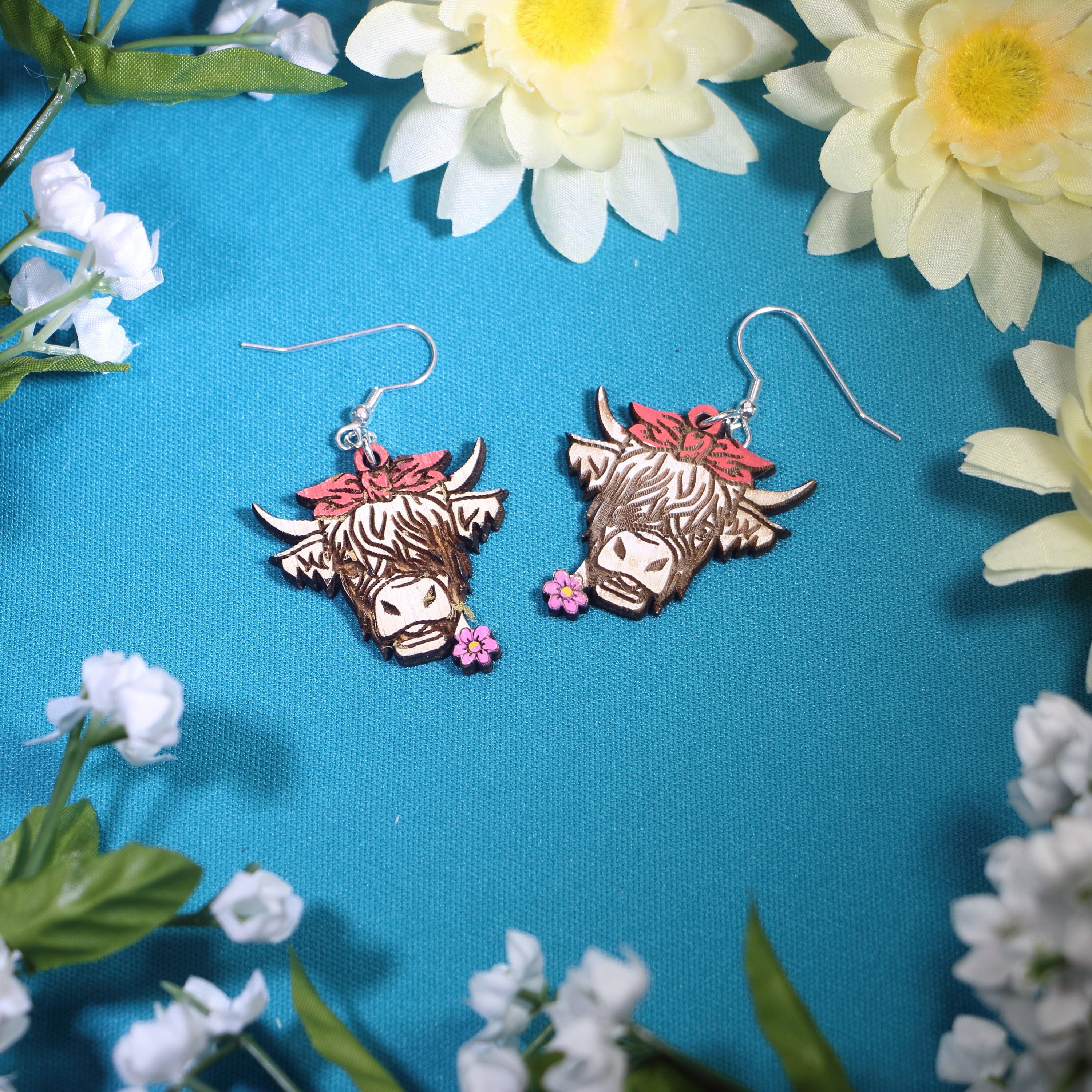 Hand-painted wooden Highland cow with flower dangle earrings featuring a shaggy-haired cow design with pink flowers and a bow, hypoallergenic sterling silver hooks, and lightweight farm animal jewelry for western style and country charm by Cate’s Concepts