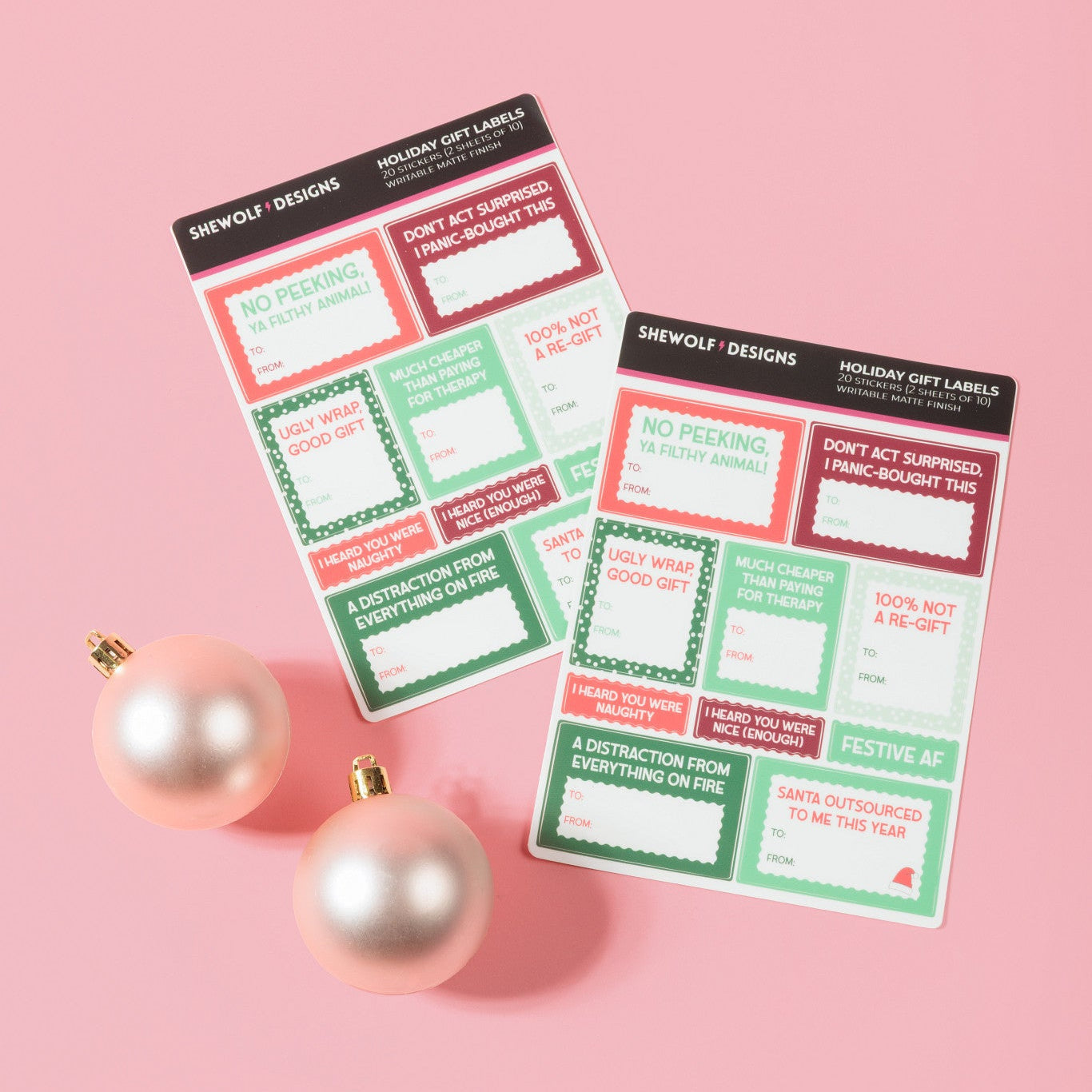 Two holiday-themed gift label sheets from SHEWOLF Designs on a pink background, styled with two pink ornaments. The notepads feature festive red, green, and white holiday labels