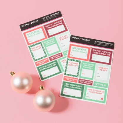 Two holiday-themed gift label sheets from SHEWOLF Designs on a pink background, styled with two pink ornaments. The notepads feature festive red, green, and white holiday labels