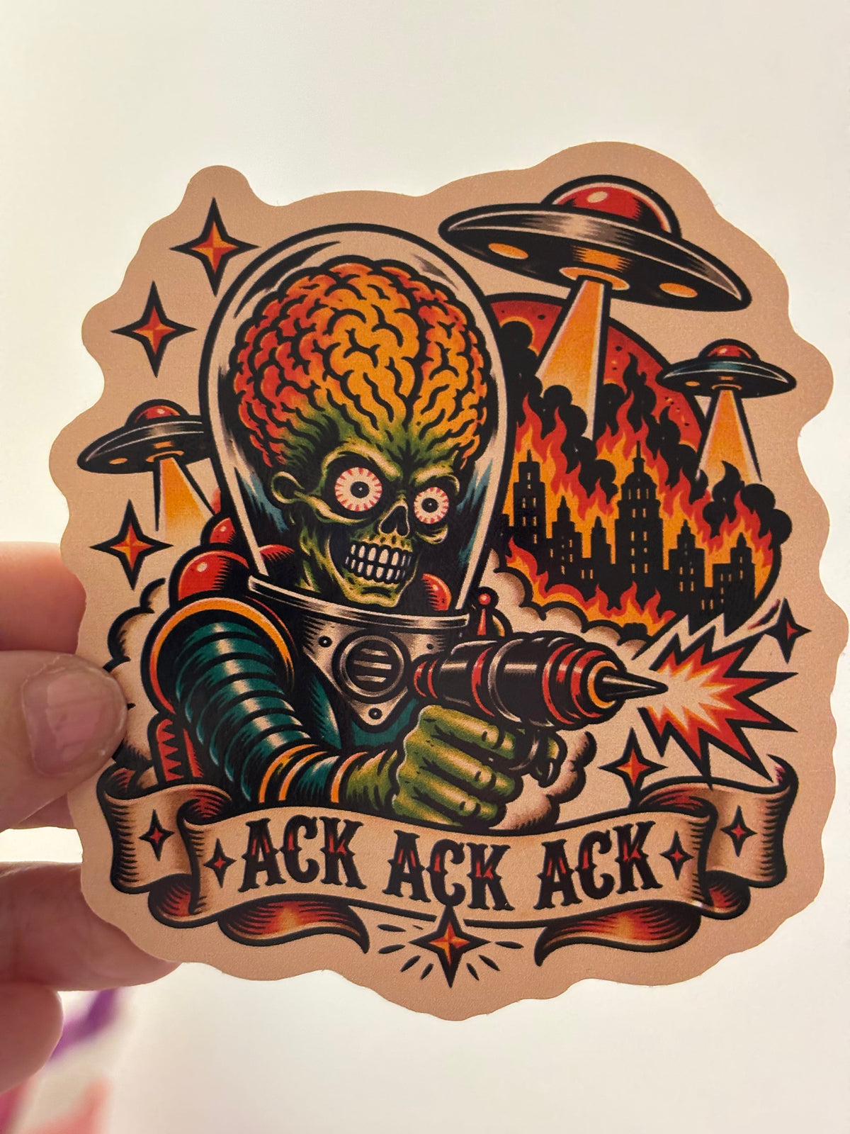 A hand holds a die-cut sticker featuring a retro-style alien-themed illustration with a green skull-headed figure in space gear firing a cannon, surrounded by UFOs, stars, and the text &