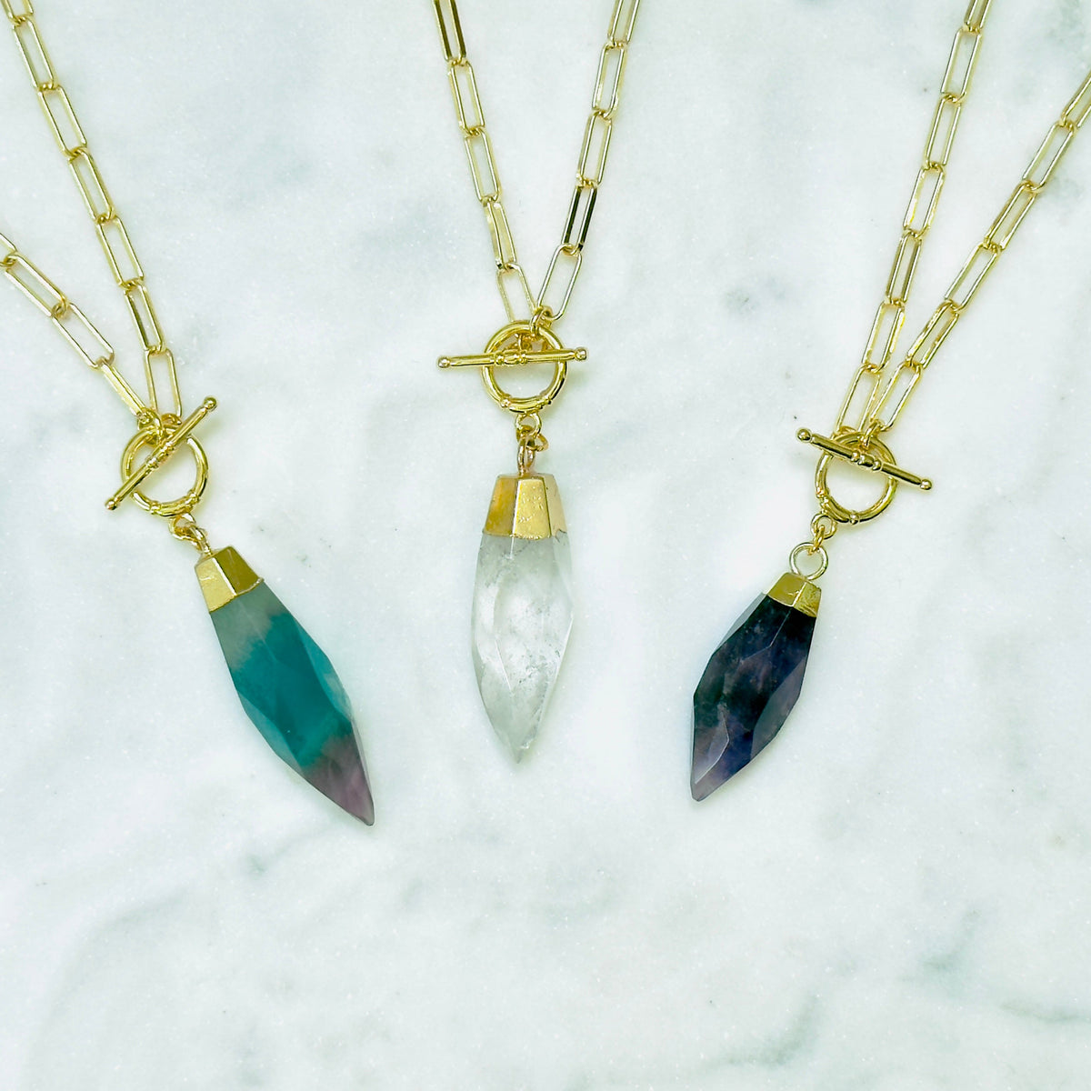 Three gold chain necklaces with toggle clamps and teardrop-shaped crystal pendants are arranged on a white marble surface. The left pendant is teal with a purple gradient at the tip, the center pendant is milky white, and the right pendant is dark blue with a purple gradient at the tip.