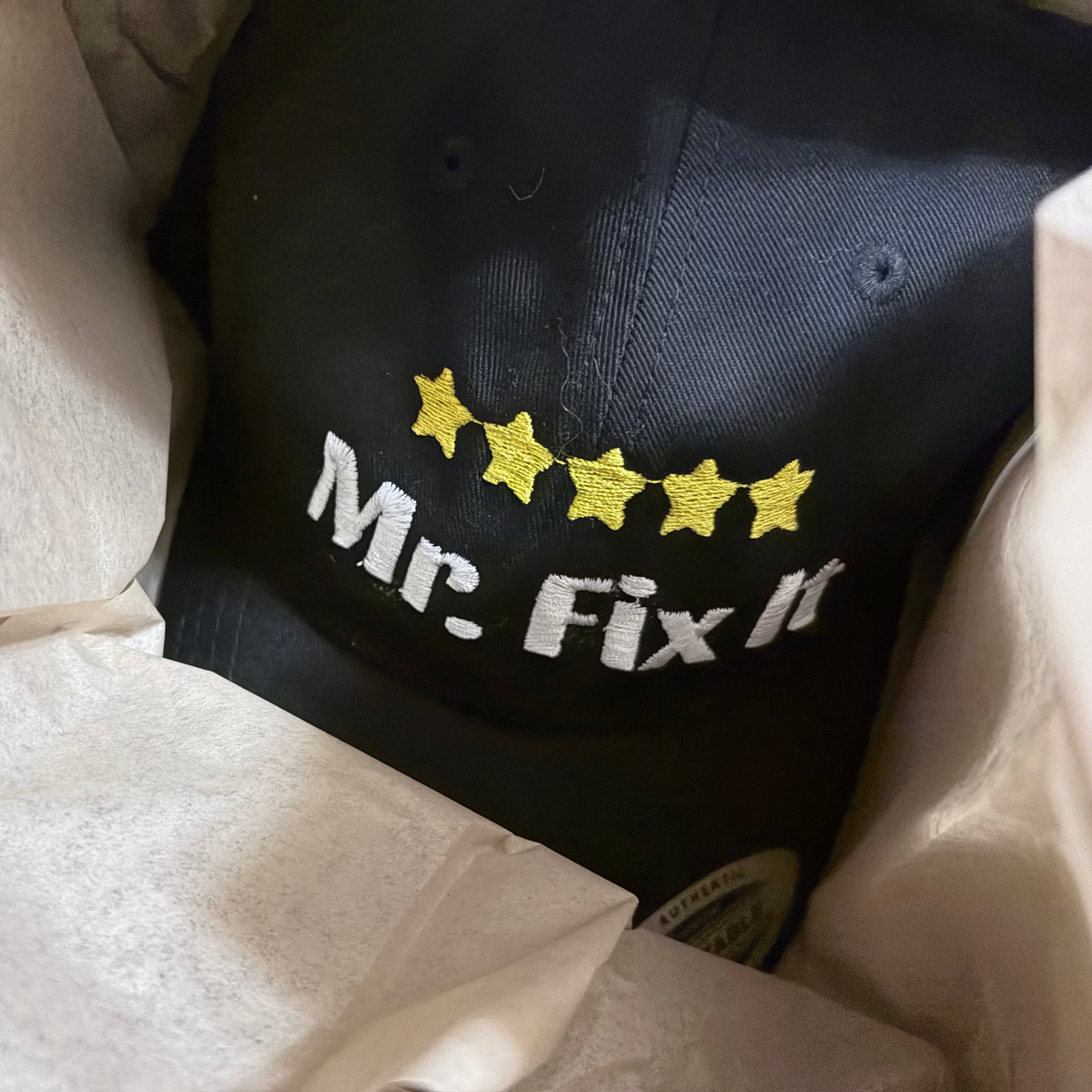 A black baseball cap nestled in white tissue paper, featuring white embroidered text reading &