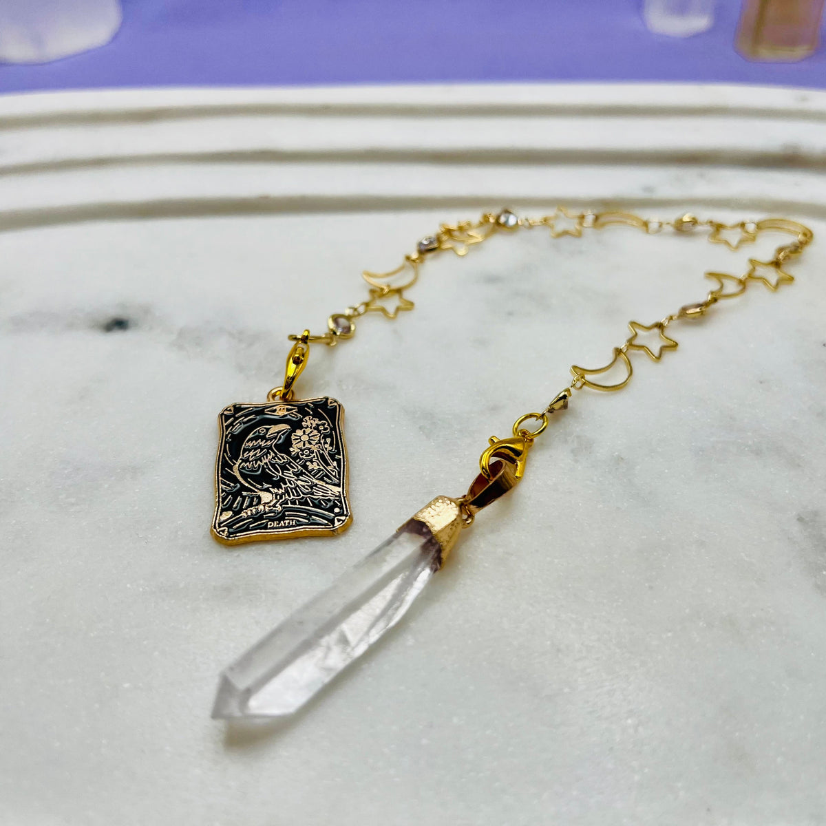 A gold-toned necklace with a rectangular engraved pendant and a clear crystal point pendant, displayed on a white marble surface.