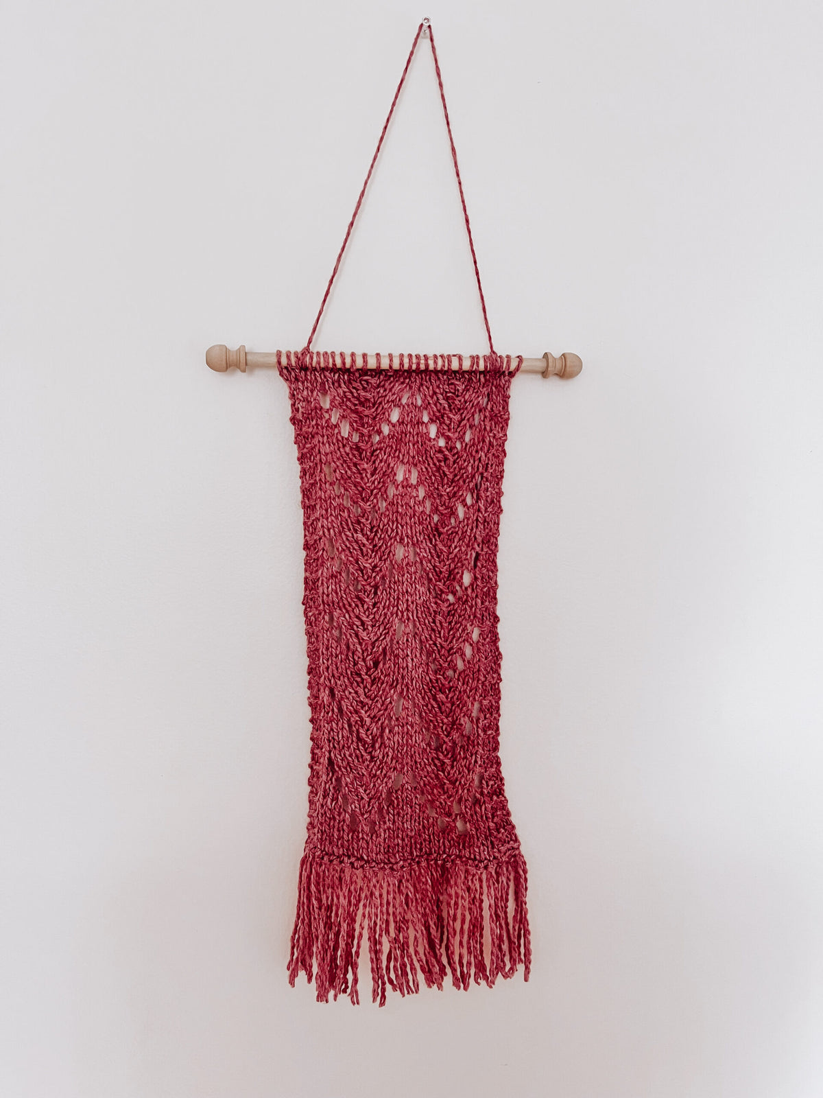 A hand dyed heathered clay color wool wall hanging is attached to a wooden dowel, hung from a longer string of wool. The hanging shows knitted chevrons in 2 columns with fringe on the bottom of the wall hanging.