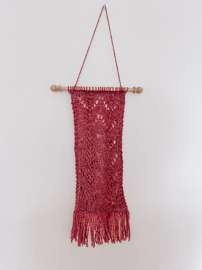 A hand dyed heathered clay color wool wall hanging is attached to a wooden dowel, hung from a longer string of wool. The hanging shows knitted chevrons in 2 columns with fringe on the bottom of the wall hanging.