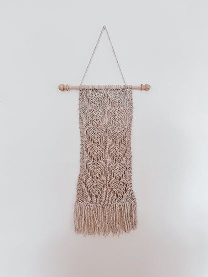 A natural tan wool wall hanging is attached to a wooden dowel, hung from a longer string of wool. The hanging shows knitted chevrons in 2 columns with fringe on the bottom of the wall hanging.