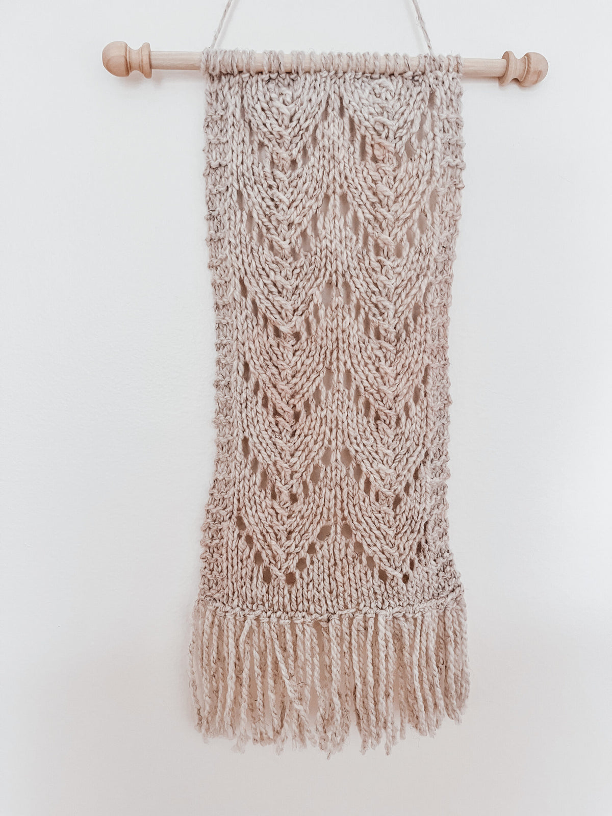 A closeup of a natural tan wool wall hanging shows knitted chevrons in 2 columns with fringe on the bottom of the wall hanging