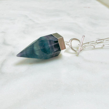 A silver pendant necklace featuring a faceted dark teal-blue gemstone point, resting on a white marble surface with a silver paperclip chain.