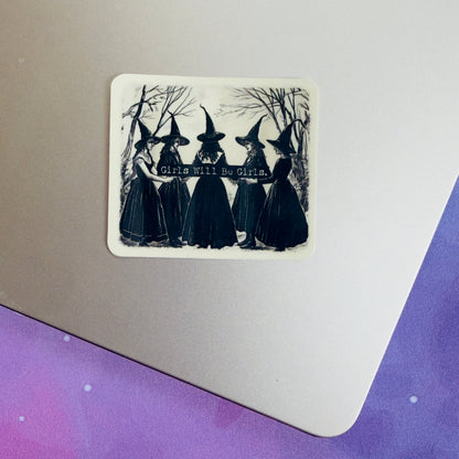 A rectangular sticker with rounded corners displayed on the corner of a silver laptop. The sticker features a black and white illustration of five witches in pointed hats standing together with bare trees in the background and the text &