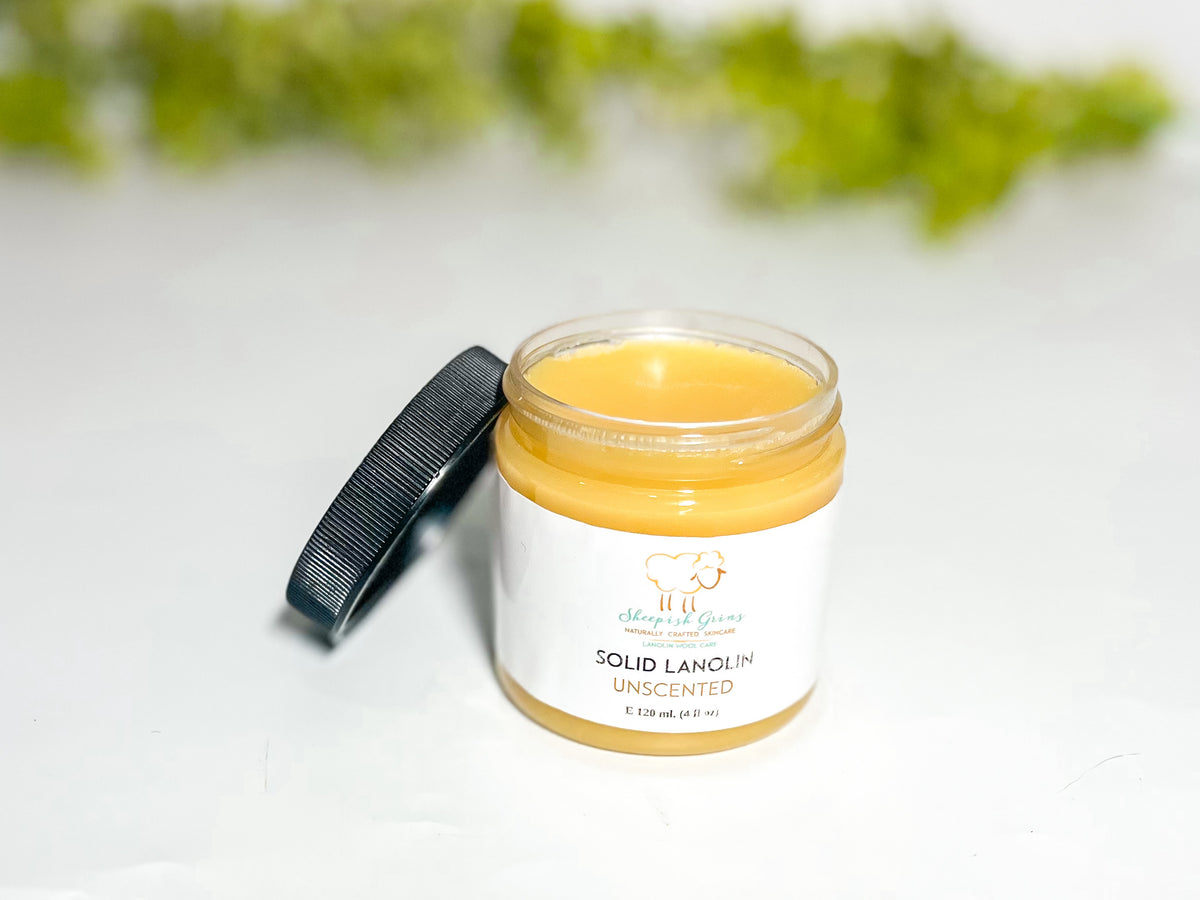 A 4 oz jar of lanolin is shown with its black lid off and leaning against the jar so the viewer can see the natural cream colored lanolin. In the background is some blurry greenery