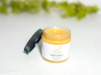 A 4 oz jar of lanolin is shown with its black lid off and leaning against the jar so the viewer can see the natural cream colored lanolin. In the background is some blurry greenery