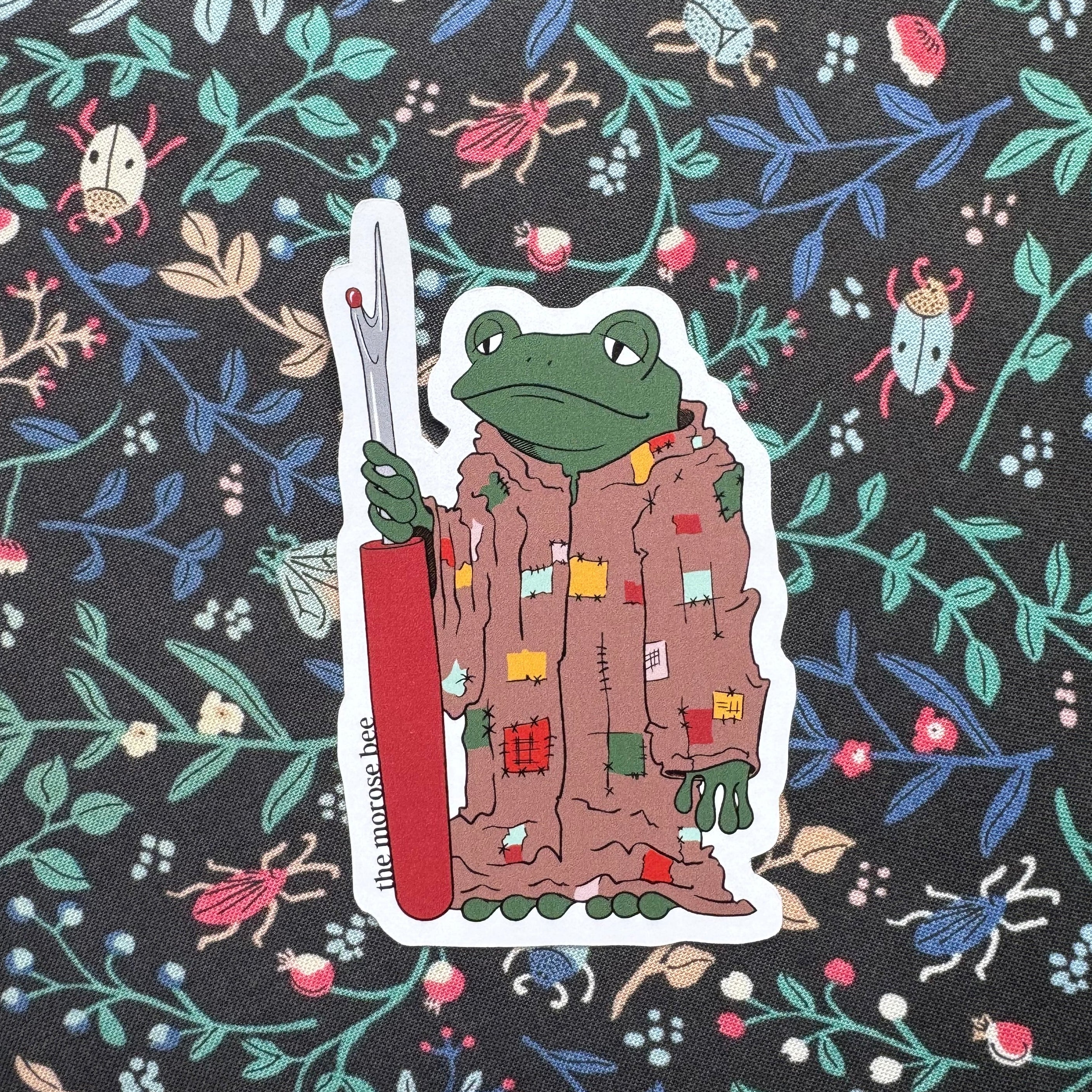 Sticker of a frog character holding a seam ripper on a floral patterned background