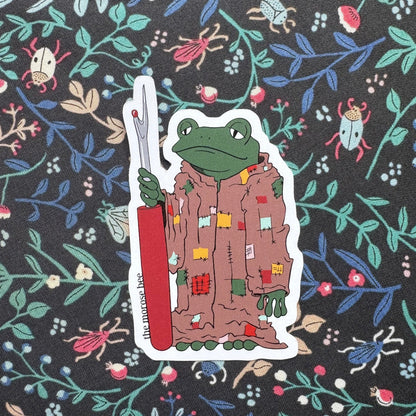 Sticker of a frog character holding a seam ripper on a floral patterned background