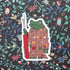 Sticker of a frog character holding a seam ripper on a floral patterned background