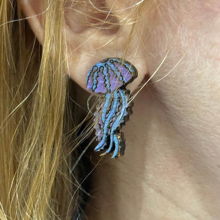 Lifestyle close-up of hand-painted wooden jellyfish dangle earrings with blue and purple ocean colors and hypoallergenic sterling silver hooks, lightweight marine life statement jewelry worn on ear