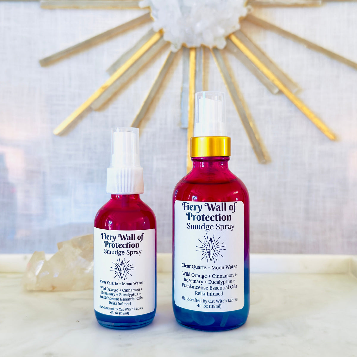 Two bottles of Firey Wall of Protection Smudge Spray, one in a red glass bottle and one in a blue glass bottle, both with white labels and spray tops, displayed on a white marble surface in front of a gold sunburst wall decor.