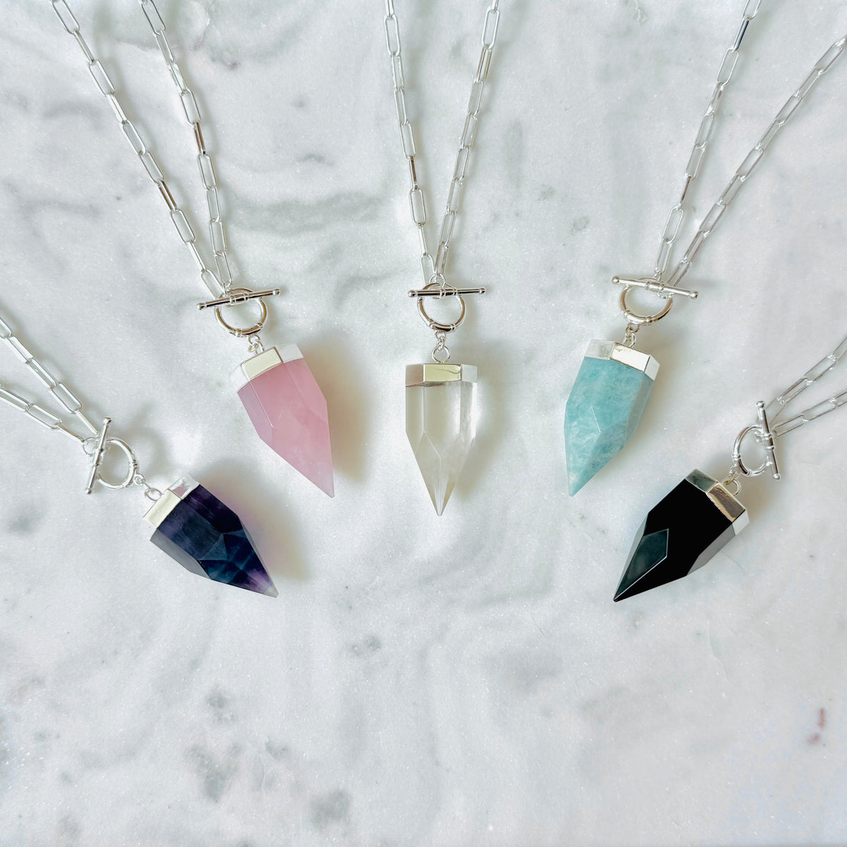 Four crystal point pendant necklaces with silver chains and toggle closures, arranged on a white marble surface. The crystals are colored dark blue, pink, clear, and black.