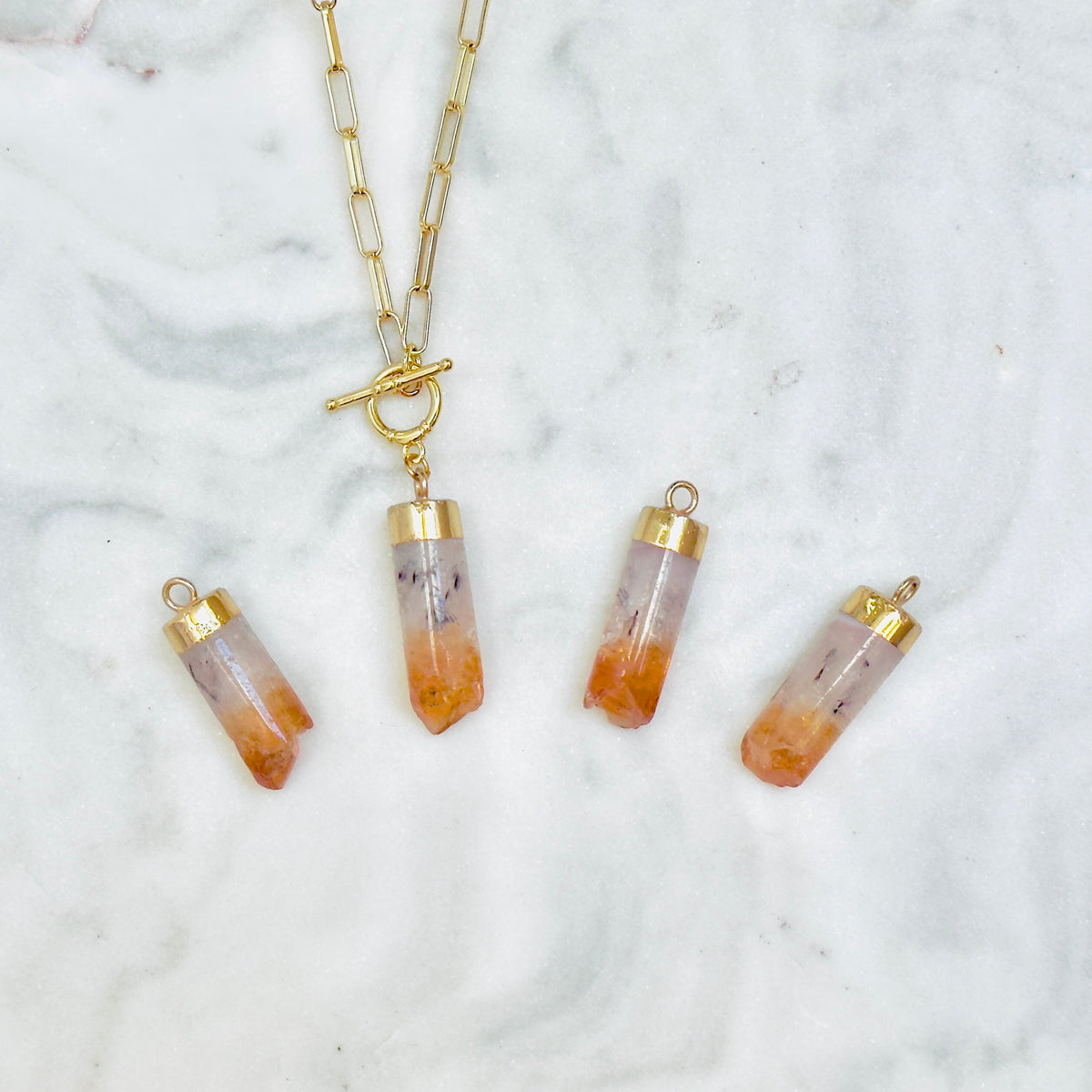 A gold chain necklace with a toggle clasp and four matching cylindrical pendant charms featuring orange and white gradient stone-like tops, arranged on a white marble surface.