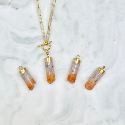 A gold chain necklace with a toggle clasp and four matching cylindrical pendant charms featuring orange and white gradient stone-like tops, arranged on a white marble surface.