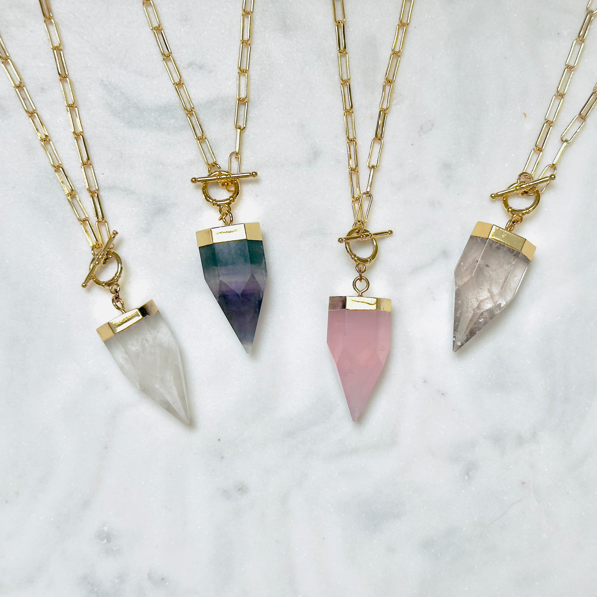 Four gold chain necklaces with toggle clasp and pointed crystal pendants in white, green-to-purple, pink, and smoky gray, arranged on a white marble surface.