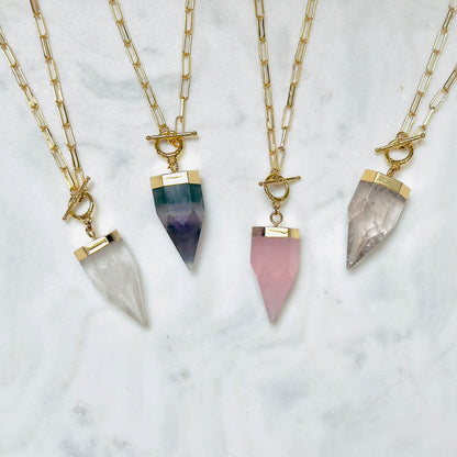 Four gold chain necklaces with toggle clasp and pointed crystal pendants in white, green-to-purple, pink, and smoky gray, arranged on a white marble surface.