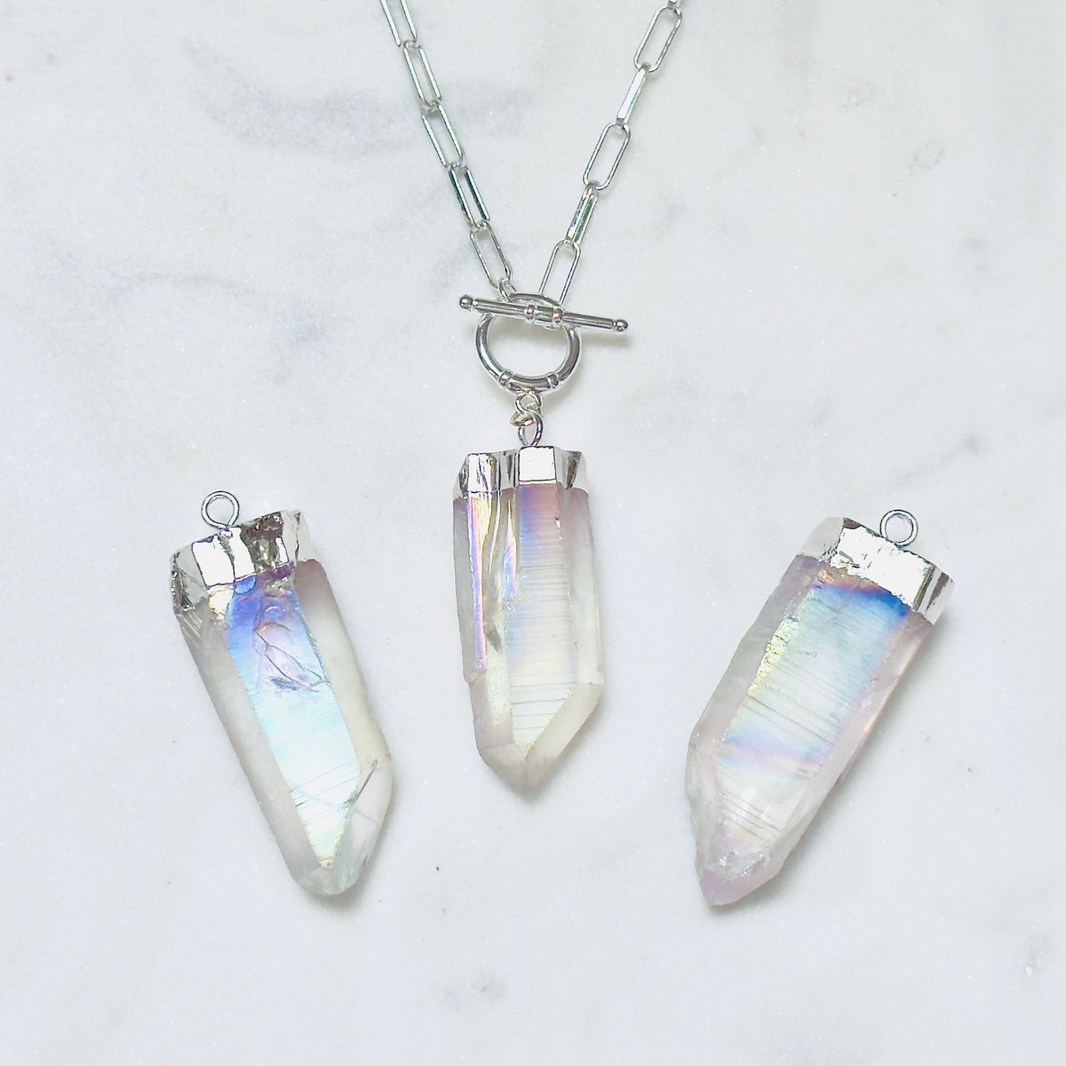 Three silver-toned crystal point pendants on a silver chain, displayed on a white marble surface. The crystals have an iridescent, milky white appearance with subtle pastel rainbow reflections.