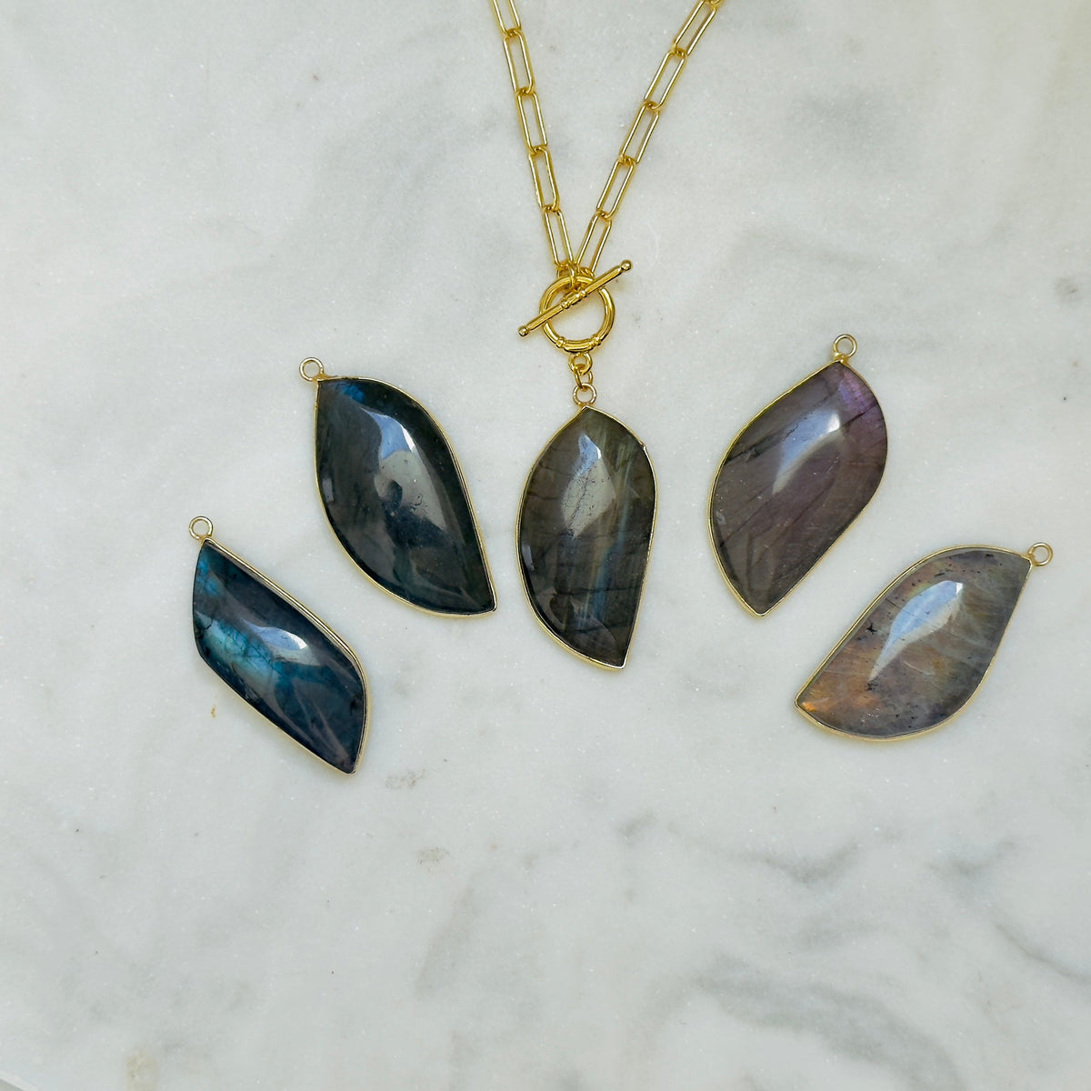 A gold chain necklace with a teardrop pendant and four matching loose teardrop pendants in different iridescent stone colors, arranged on a white marble surface.