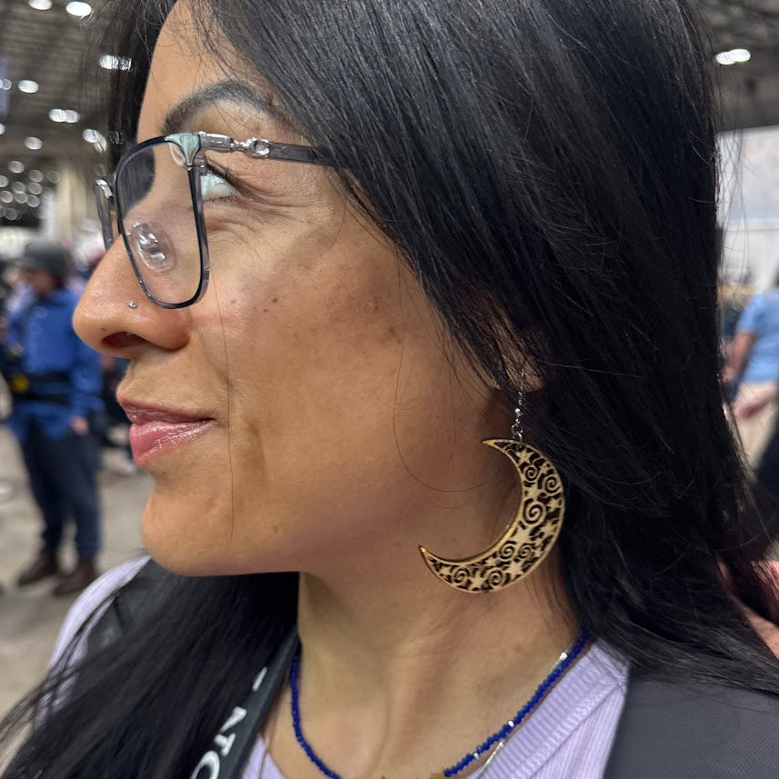 Lifestyle photo of woman wearing wooden crescent moon dangle earrings with intricate filigree cutout design and hypoallergenic sterling silver hooks, celestial boho statement jewelry at indoor market