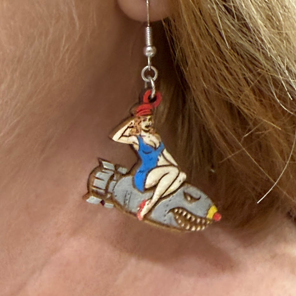 Close-up of hand-painted wooden pin-up bomber girl dangle earrings featuring vintage nose art style woman riding cartoon bomb with red hat and blue outfit and hypoallergenic sterling silver hooks, retro WWII inspired statement jewelry worn on ear