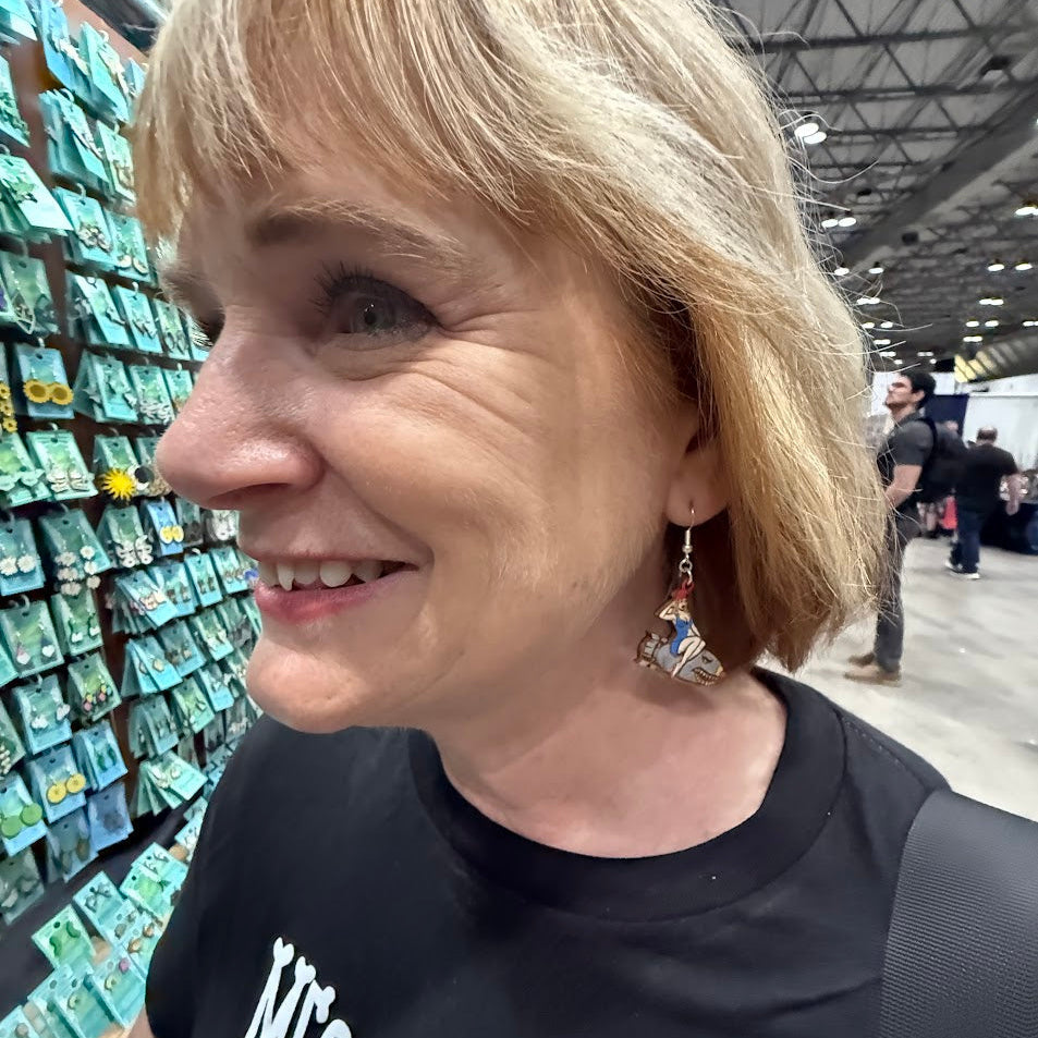 Lifestyle photo of woman wearing hand-painted wooden pin-up bomber girl dangle earrings featuring vintage nose art style woman riding cartoon bomb with red hat and blue outfit and hypoallergenic sterling silver hooks, retro WWII inspired statement jewelry at planet comic con
