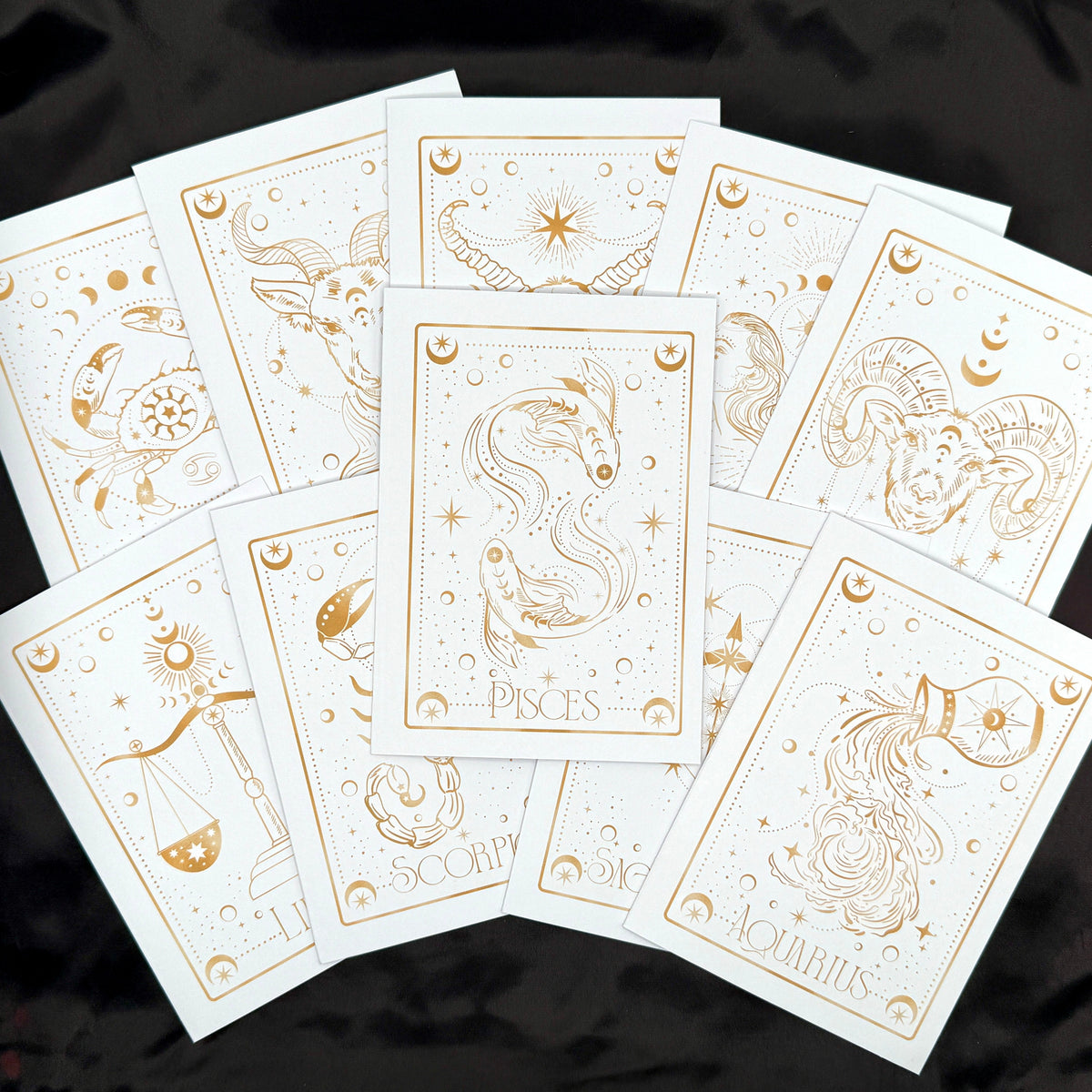 A fan arrangement of white zodiac tarot cards with elegant gold line art and titles including Pisces, Scorpio, Aquarius, and others, displayed on a black fabric background.