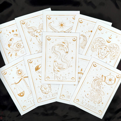 A fan arrangement of white zodiac tarot cards with elegant gold line art and titles including Pisces, Scorpio, Aquarius, and others, displayed on a black fabric background.