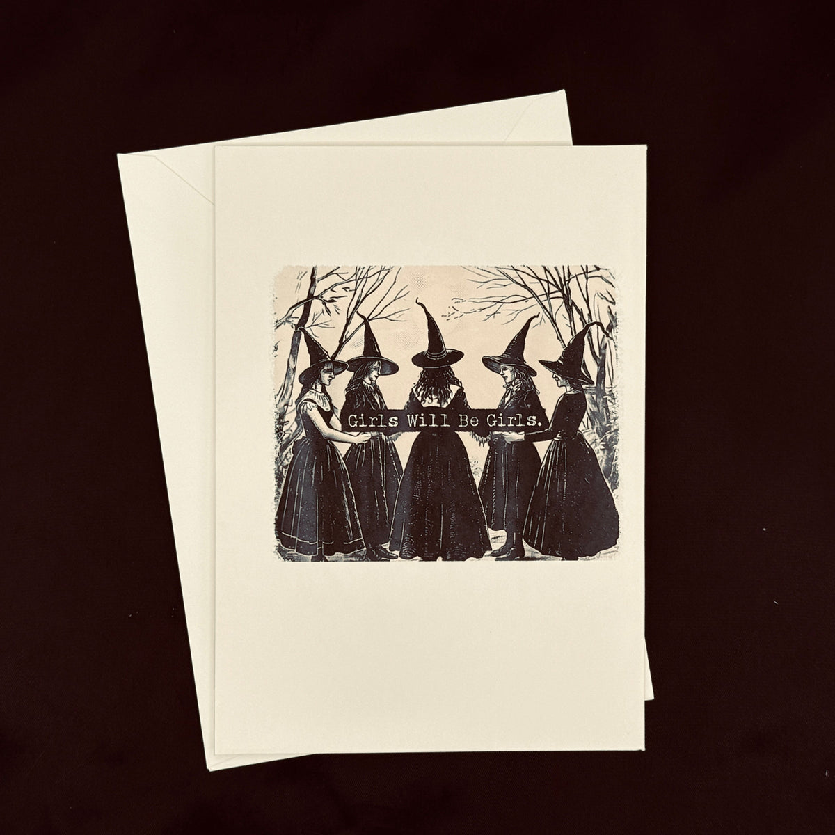 A cream-colored greeting card with a black vintage-style illustration of five witches holding a banner that reads &