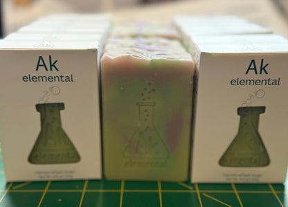 Soaps in white boxes on either side of a pale green soap stamped with the AK Elemental logo.