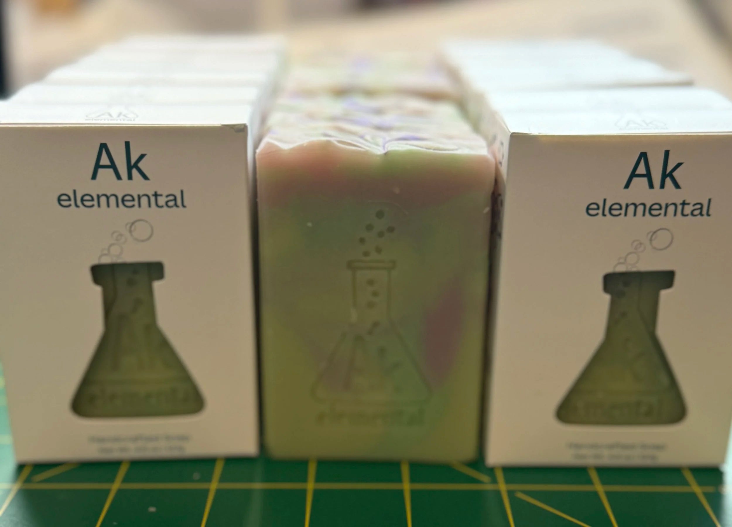 Soaps in white boxes on either side of a pale green soap stamped with the AK Elemental logo.