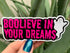 Artist holding a glossy pink and black sticker reading boolieve in your dreams and featuring a smiling ghost