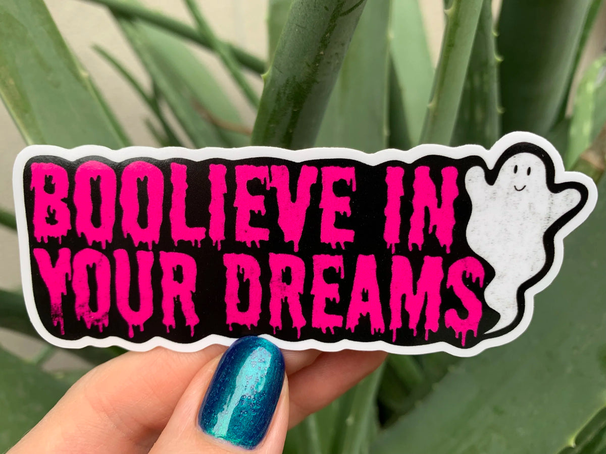 Artist holding a glossy pink and black sticker reading boolieve in your dreams and featuring a smiling ghost