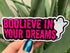 Artist holding a glossy pink and black sticker reading boolieve in your dreams and featuring a smiling ghost