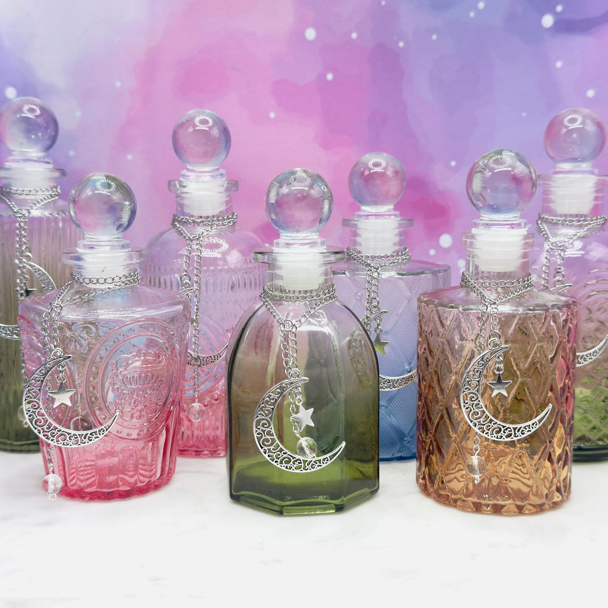 A set of pastel gradient glass perfume bottles with silver crescent moon and star designs, arranged on a white marble surface against a pink and purple galaxy backdrop.