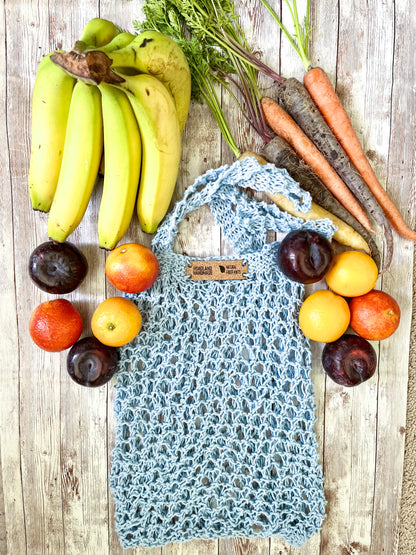 A light blue recycled cotton mesh tote bag is surrounded by bananas, rainbow carrots, plums, oranges, and blood oranges