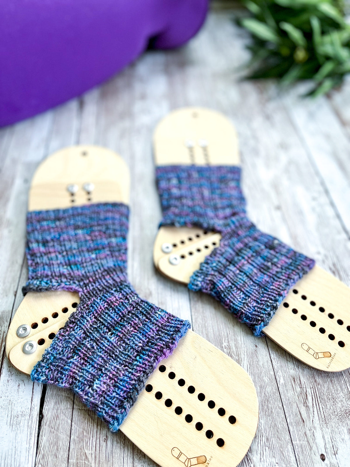 A pair of purple and blue yoga socks on wooden sock blockers are surrounded by a rolled up yoga mat and greenery on a wood panel