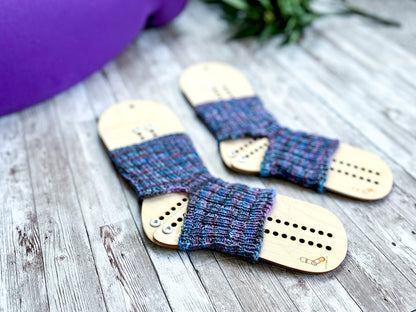 A pair of purple and blue yoga socks on wooden sock blockers are surrounded by a rolled up yoga mat and greenery on a wood panel
