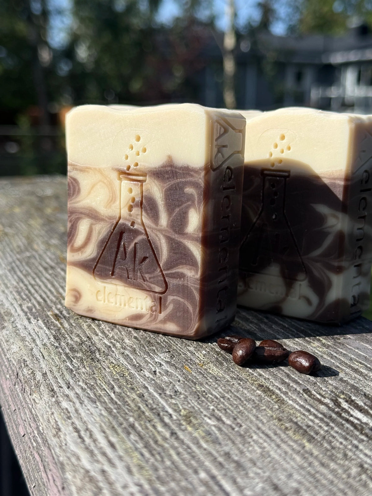 Three bars of soap with brown and cream swirls and cream top sitting on a weathered board. AK Elemental logo of an erlenmeyer flask is stamped on the soaps.