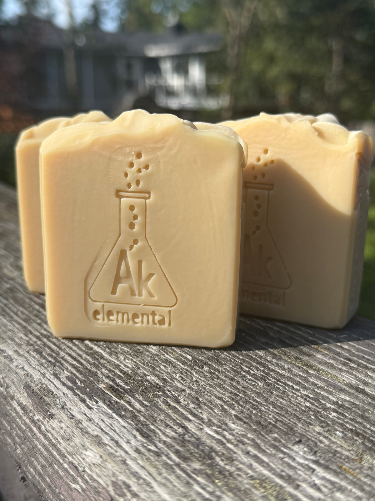 Three creamy, uncolored Sugar Cookie handcrafted soap bars with an embossed AK Elemental flask logo, displayed on rustic wood outdoors.
