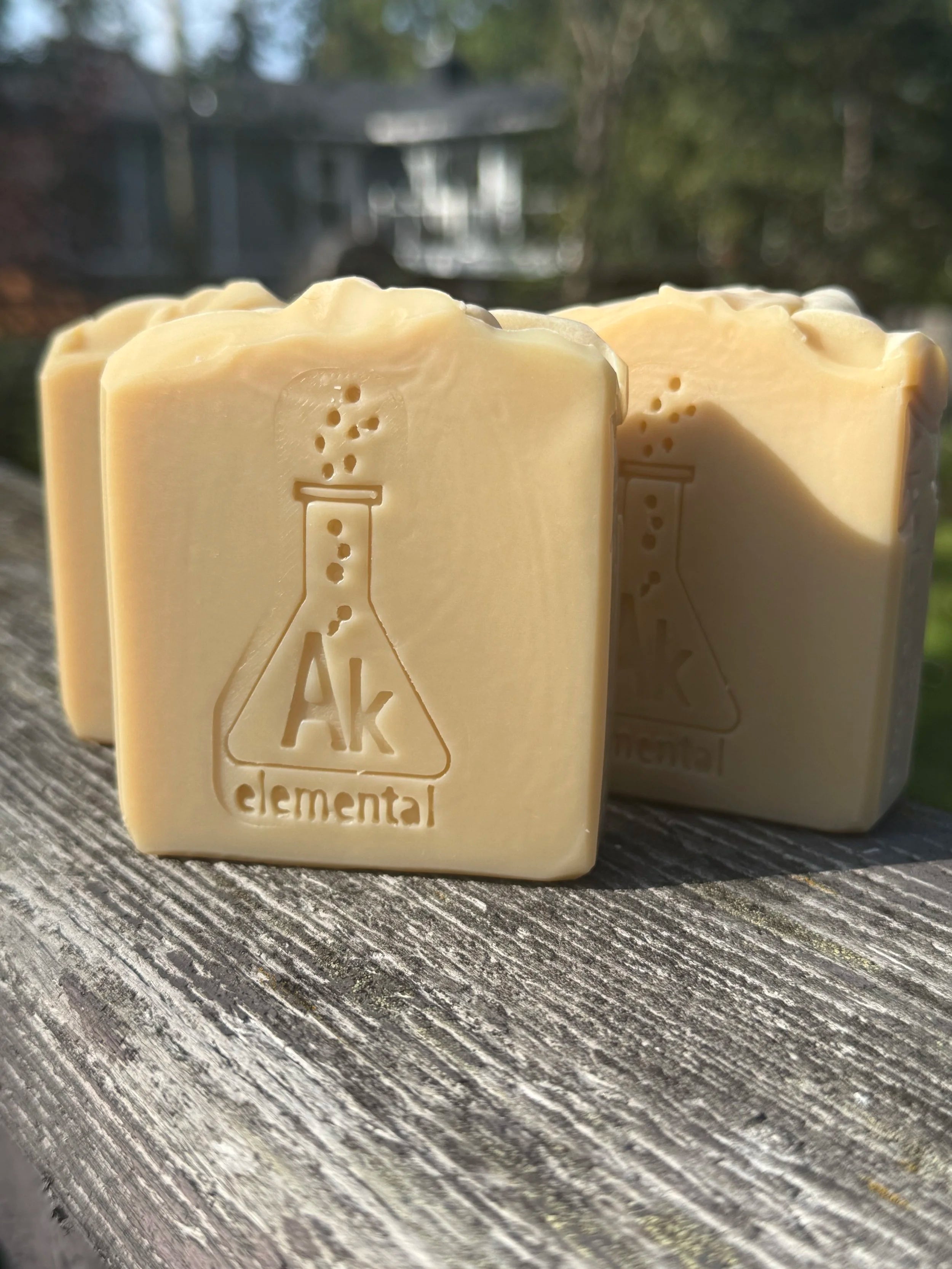 Three creamy, uncolored Sugar Cookie handcrafted soap bars with an embossed AK Elemental flask logo, displayed on rustic wood outdoors.