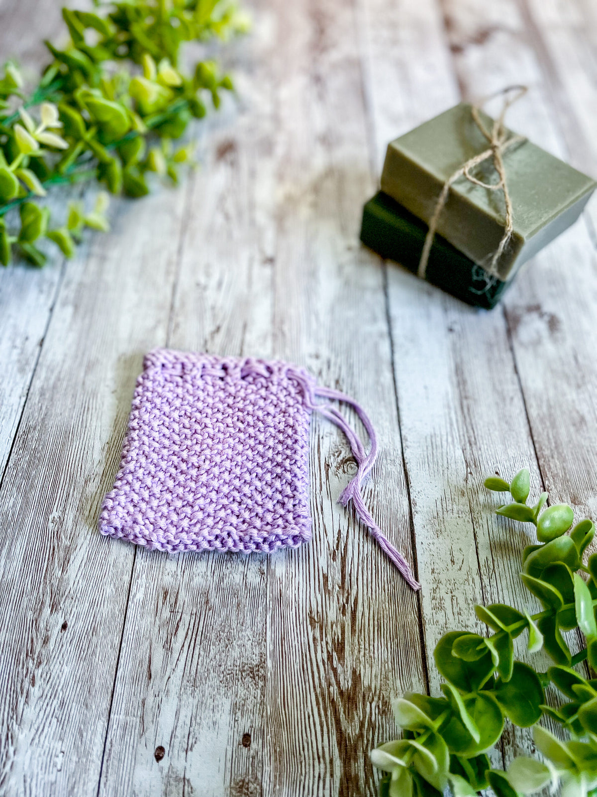 A purple bamboo/cotton soap saver rests on a wood plank next to 2 bars of soap wrapped with twine and a sprig of eucalyptus leaves