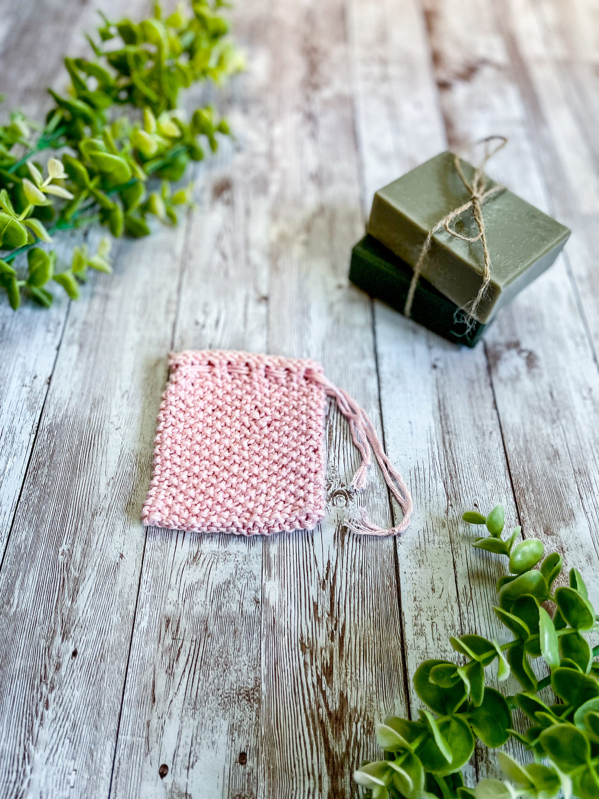 A pink bamboo/cotton soap saver rests on a wood plank next to 2 bars of soap wrapped with twine and a sprig of eucalyptus leaves