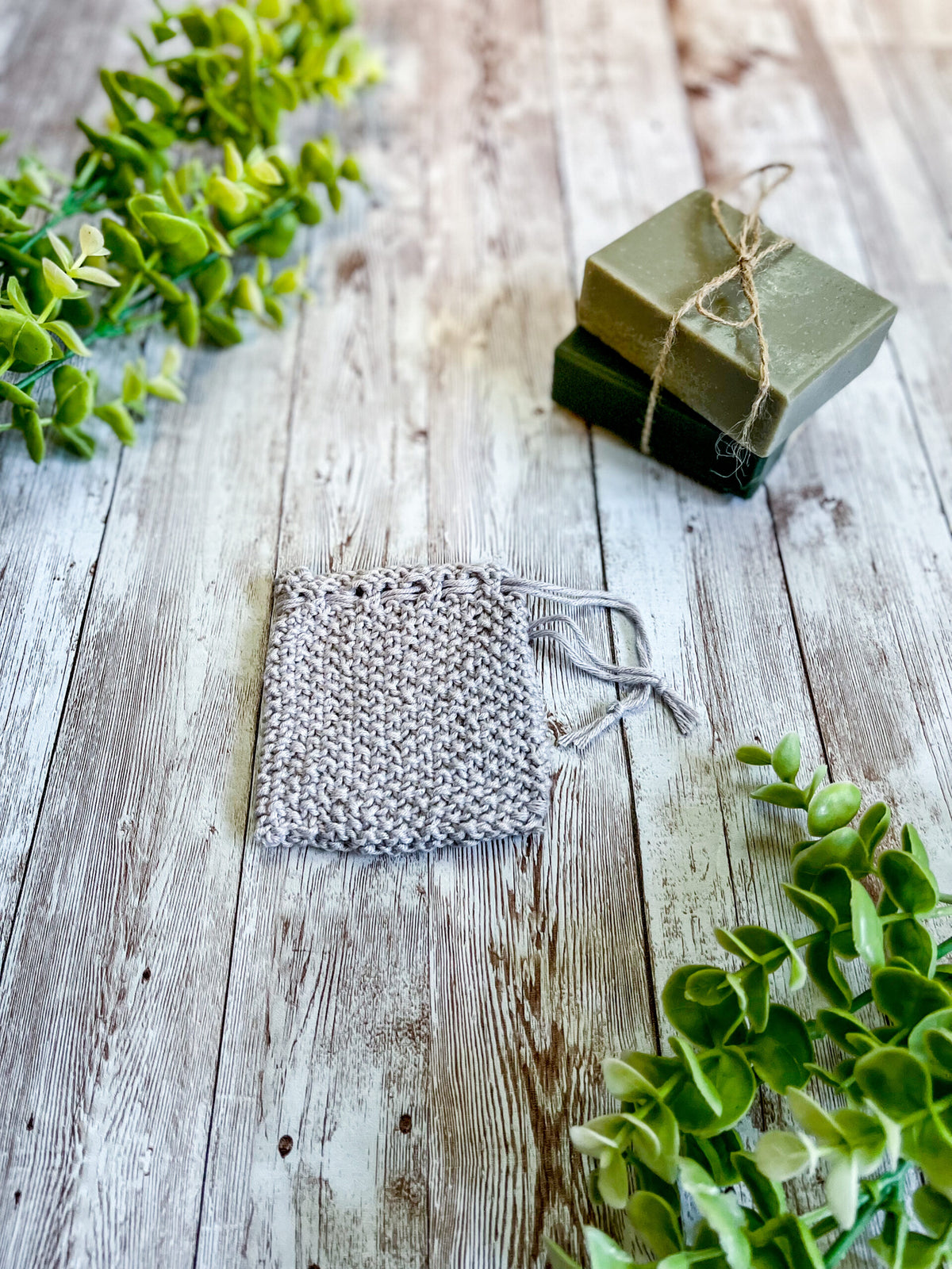 A gray bamboo/cotton soap saver rests on a wood plank next to 2 bars of soap wrapped with twine and a sprig of eucalyptus leaves